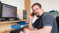 man in t shirt met sikje en bril aan de telefoon die iets opschrijft, zittend voor een computer