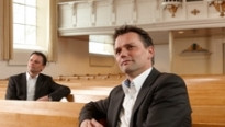 Twee mannen in pak die op kerkbanken zitten in een lege kerk met een wit balkon